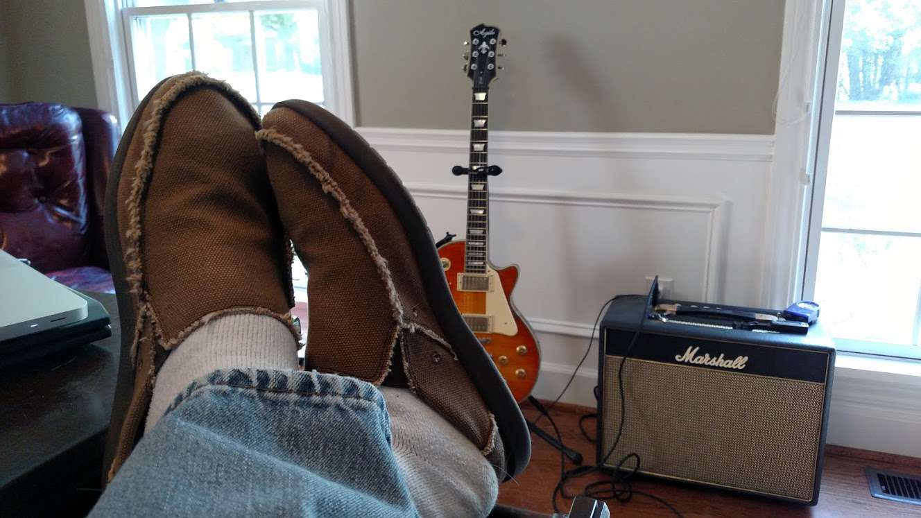 Photo of my feet up on desk near laptops with guitar and amp in background.